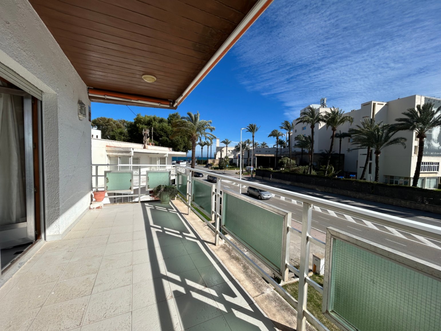 Terraza exterior con suelo cerámico, techo revestido de madera y barandilla metálica con paneles de vidrio. Ofrece vistas despejadas a la avenida y palmeras.