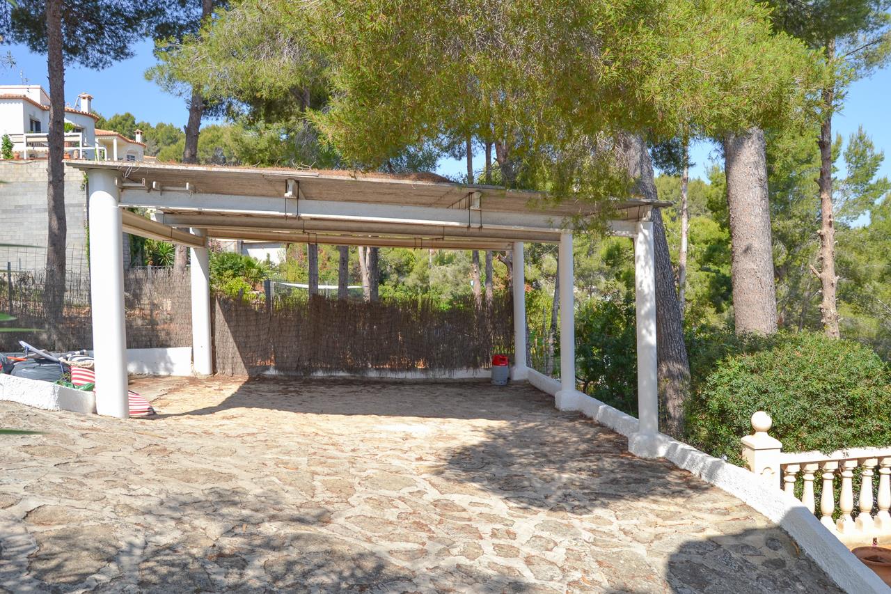 Área de aparcamiento cubierta con pilares blancos y suelo de piedra natural, rodeada de pinos y vegetación mediterránea en una zona tranquila.