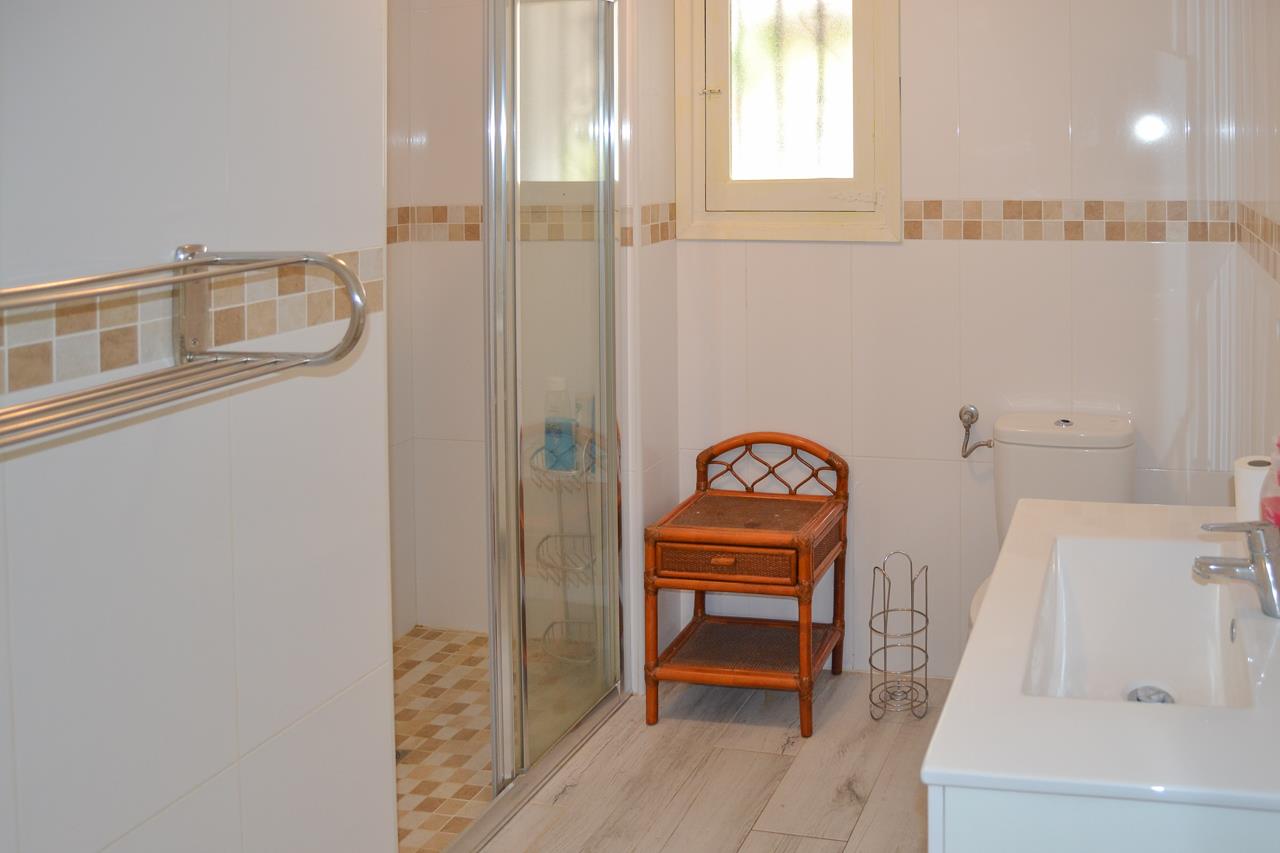 Baño con paredes alicatadas en blanco y cenefa de mosaico. Dispone de cabina de ducha con mampara de cristal, lavabo integrado y mobiliario auxiliar de mimbre.