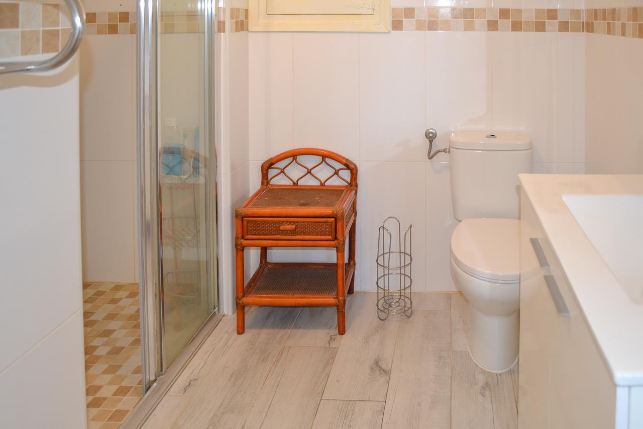 Baño funcional con ducha de mampara, inodoro blanco y mueble auxiliar de ratán. Destaca el alicatado claro con cenefa de mosaico y el suelo de gres con acabado efecto madera.