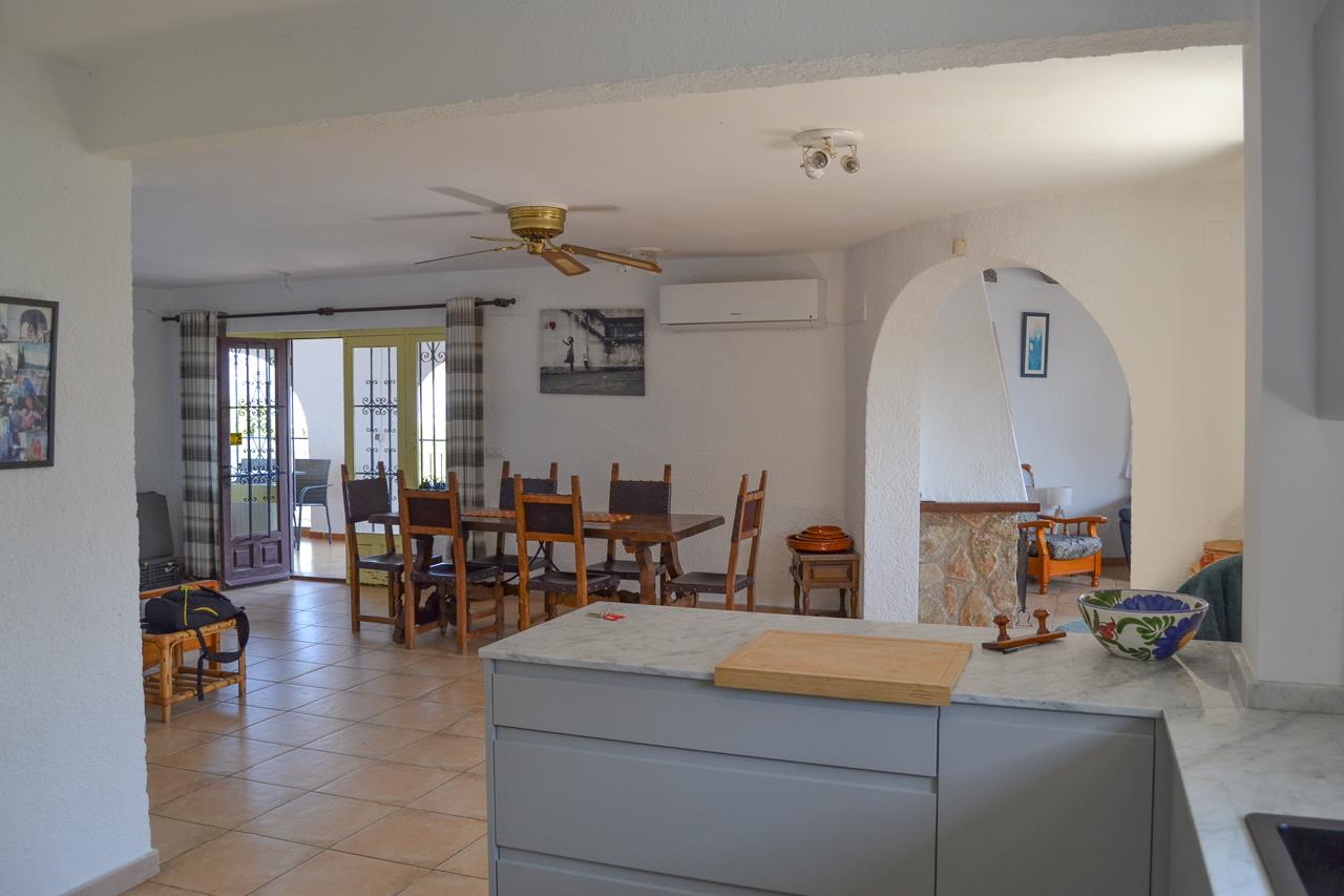 Cocina abierta con encimera de mármol integrada en un comedor luminoso. Dispone de aire acondicionado, ventilador de techo y acceso a la naya a través de ventanales.
