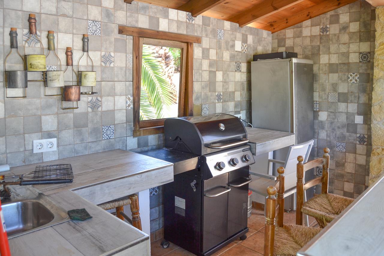 Cocina de verano techada con barbacoa de gas, encimeras de gres y paredes revestidas de azulejos rústicos. Incluye vigas de madera, fregadero y vistas a la vegetación exterior.