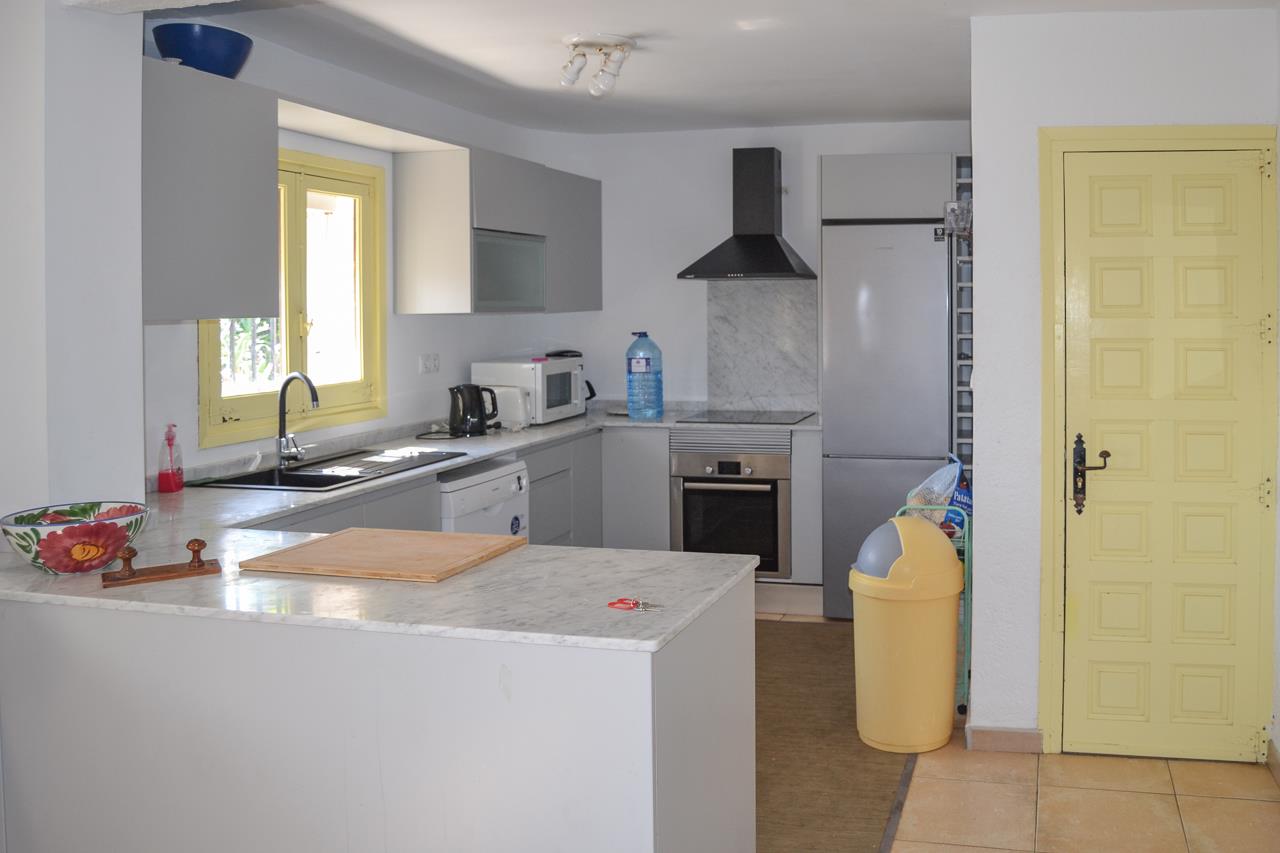 Cocina funcional con encimeras de mármol, armarios en gris suave y electrodomésticos integrados. Destaca su ventana y puerta en tono amarillo que aportan luminosidad al espacio.