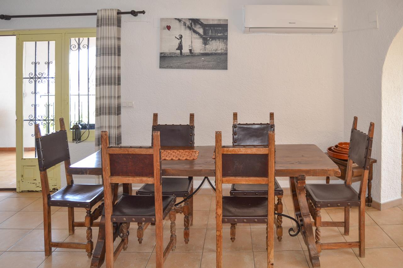 Comedor con mesa de madera maciza y sillas de cuero de estilo rústico. Dispone de aire acondicionado split y acceso directo a través de una puerta acristalada.