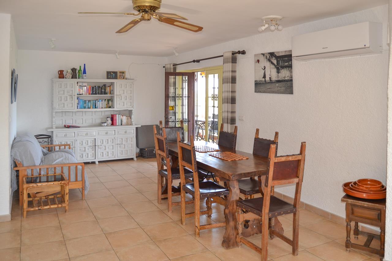 Comedor de estilo rústico con mesa de madera maciza, sillas de respaldo alto y aparador blanco. Dispone de aire acondicionado, ventilador de techo y acceso directo a la terraza.