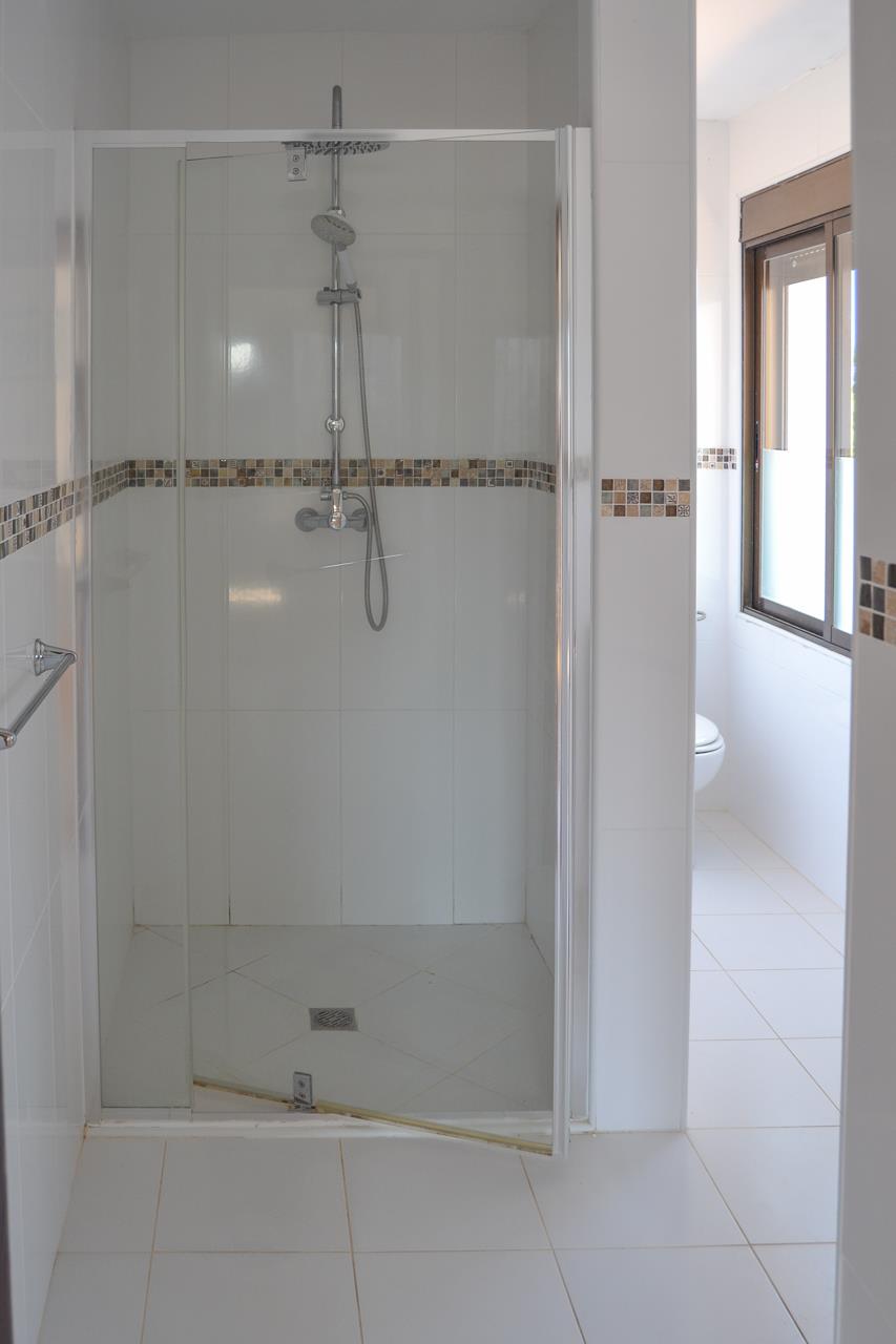 Cuarto de baño con ducha de obra, mampara de cristal, azulejos blancos y cenefa decorativa de mosaico en tonos tierra.