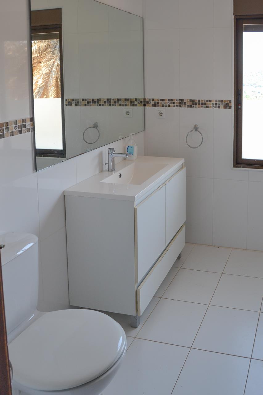 Cuarto de baño con mueble lavabo blanco, espejo de gran formato y azulejos con cenefa decorativa en tonos tierra. Incluye ventana con luz natural.