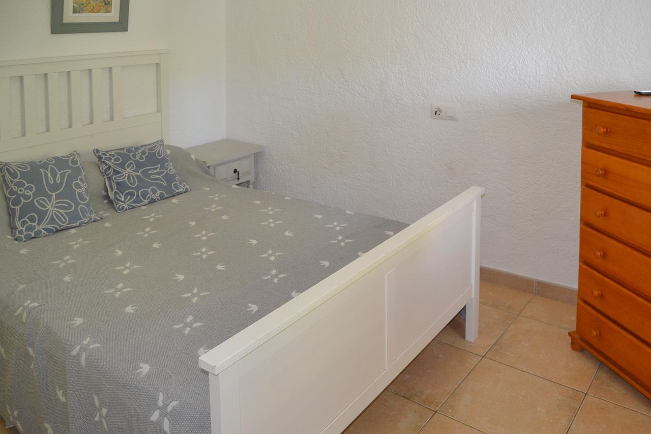 Dormitorio con cama de madera blanca, suelos de gres cerámico y una cómoda de madera natural. Las paredes blancas texturizadas aportan luminosidad al espacio.
