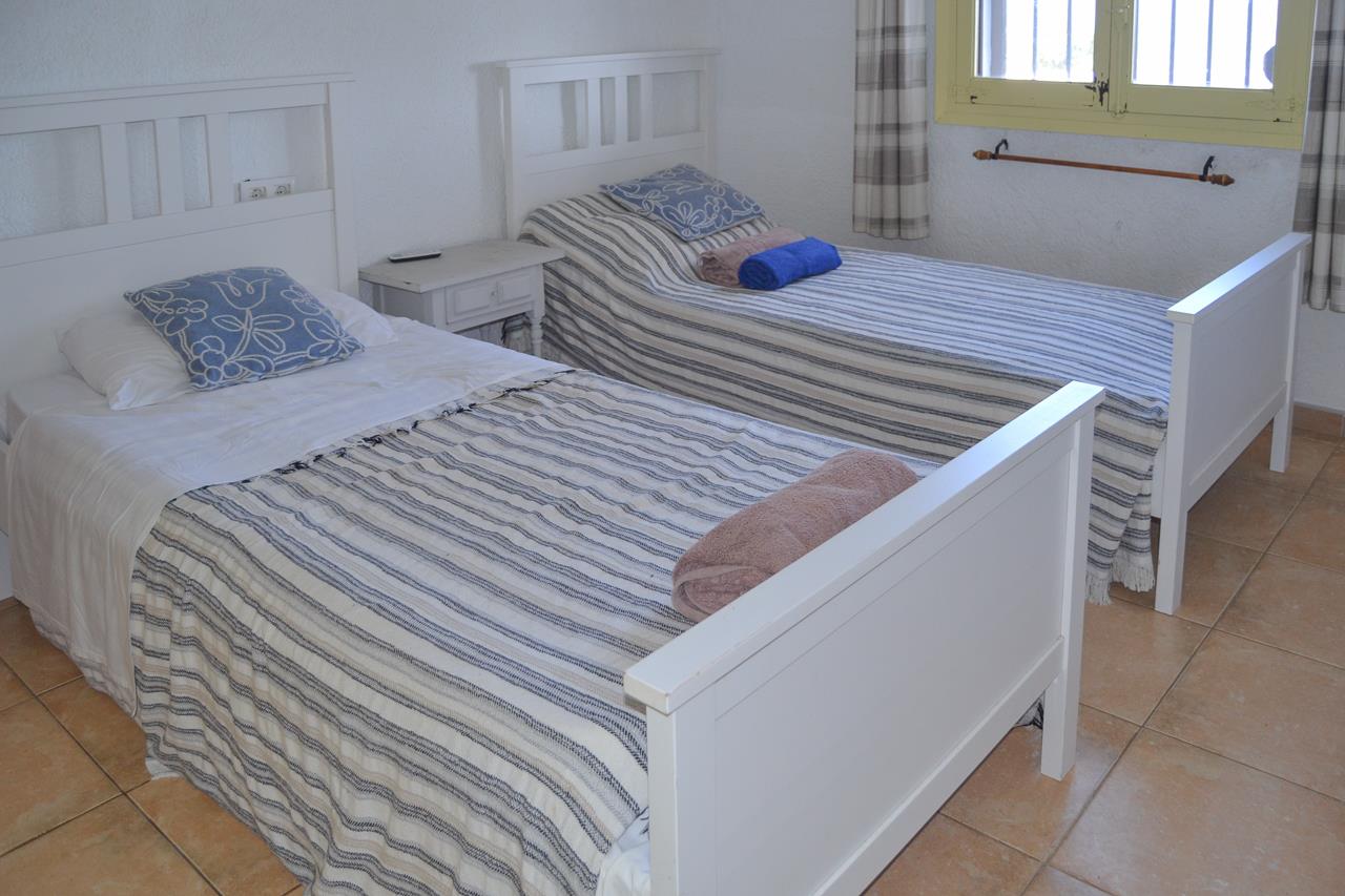 Dormitorio con dos camas individuales de madera blanca, suelos de gres rústico y ventana con marcos amarillos y cortinas de cuadros.