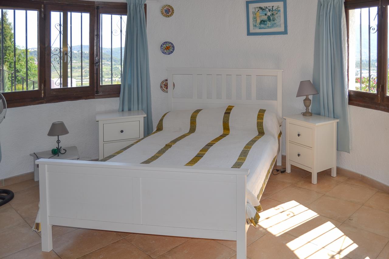Dormitorio luminoso con cama de madera blanca, suelos de terracota y ventanas con marcos de madera que ofrecen vistas despejadas al valle de Monte Pedreguer.