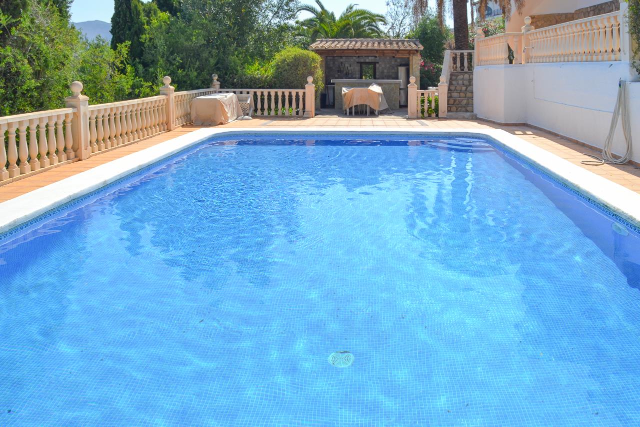 Piscina rectangular de gresite azul rodeada de una amplia terraza con balaustrada y zona de bar cubierta al fondo entre vegetación mediterránea.
