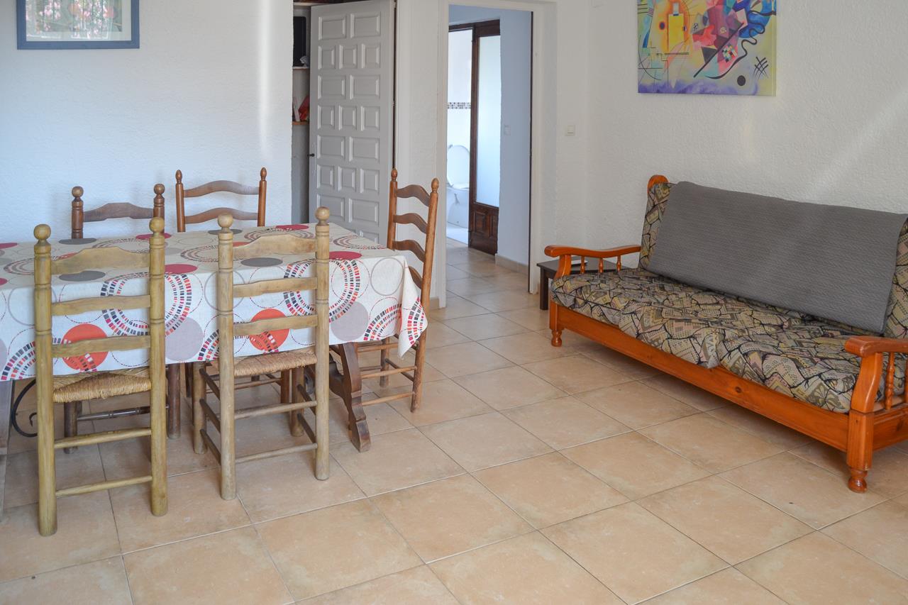 Salón comedor con suelos de gres cerámico, mesa de madera con sillas de anea y un sofá rústico. El espacio conecta directamente con el pasillo y el baño de la vivienda.
