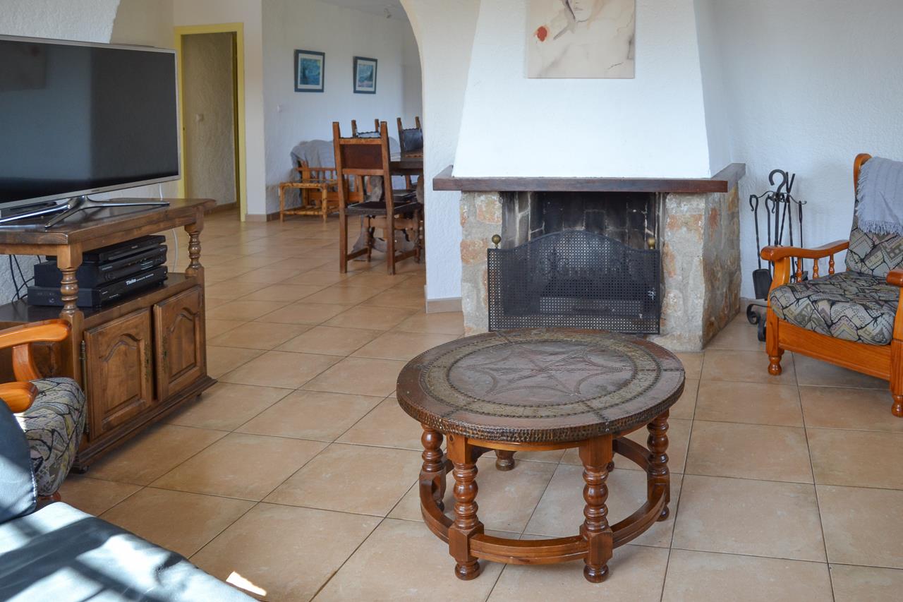 Salón rústico con chimenea de piedra, suelos de gres y mobiliario de madera tradicional. Un arco conecta el espacio de estar con el comedor adyacente.
