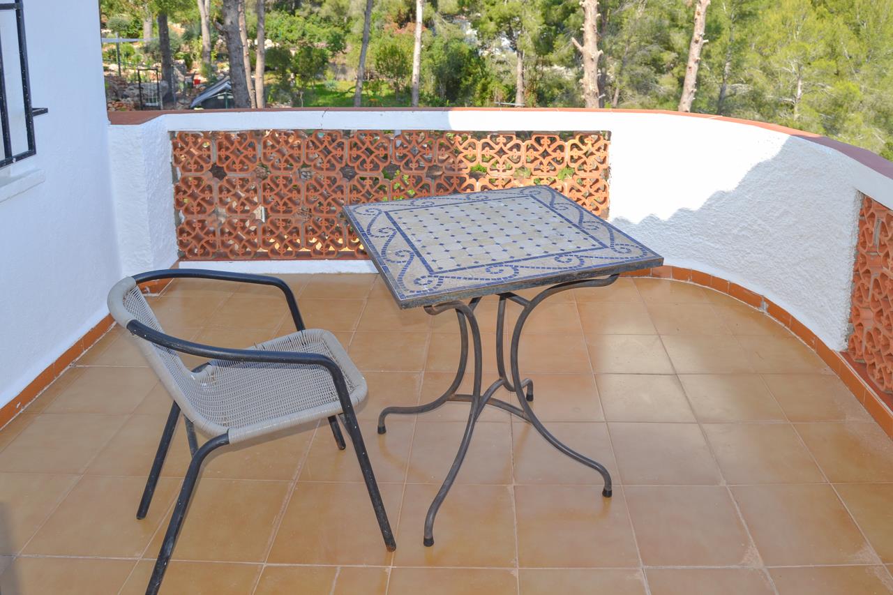Terraza exterior con suelo de barro, mesa de mosaico azul y barandilla con celosía cerámica decorativa frente a un entorno de pinos.