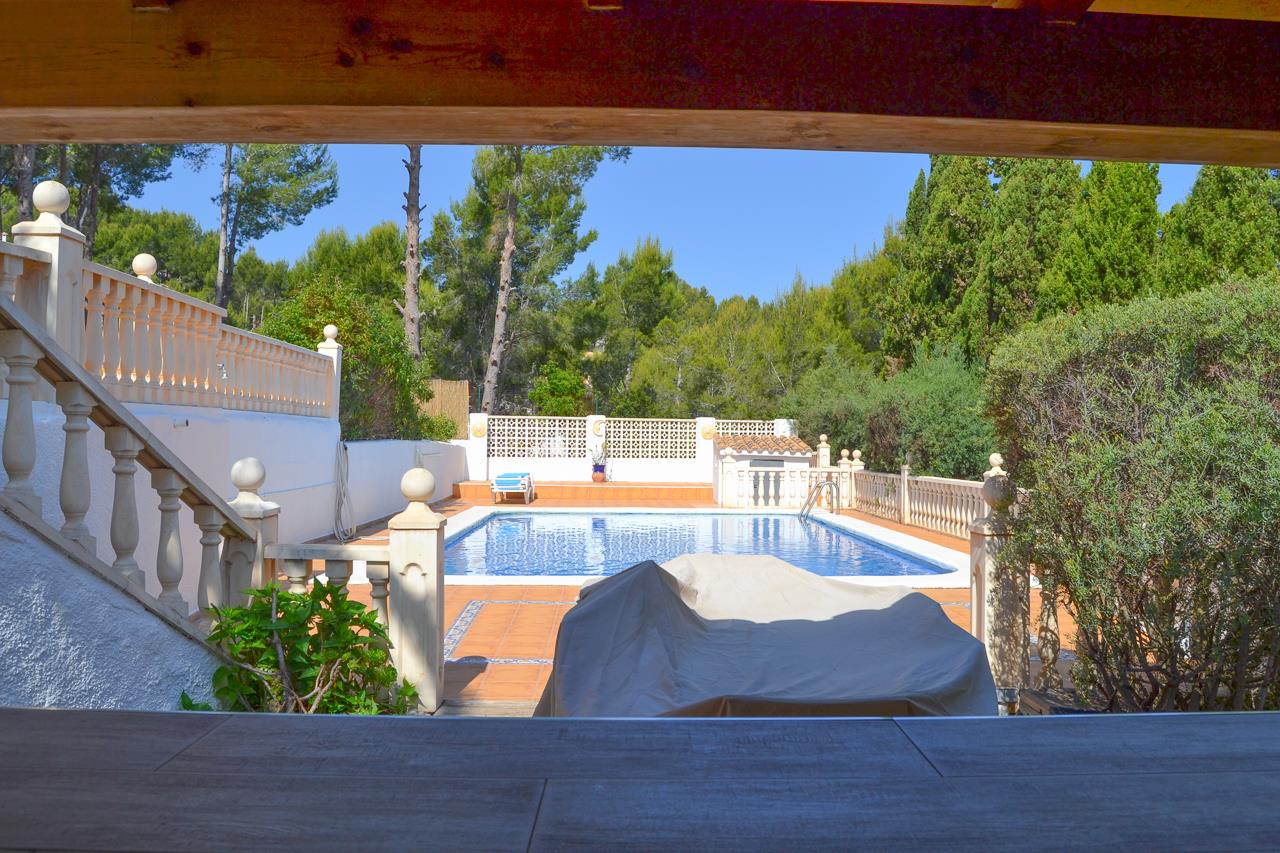Vista de la piscina de 12x6 metros desde la zona de barra cubierta, rodeada de amplias terrazas soleadas, balaustradas de piedra y vegetación mediterránea.