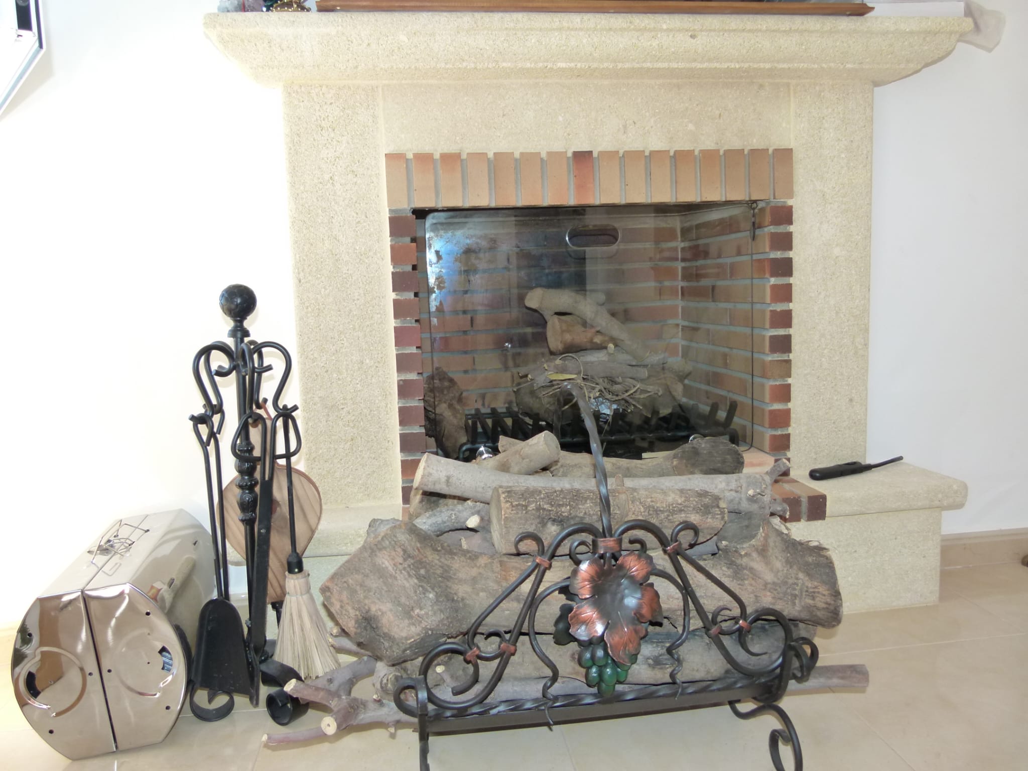 Chimenea de ladrillo con accesorios de hierro forjado y leña apilada sobre soporte decorativo.