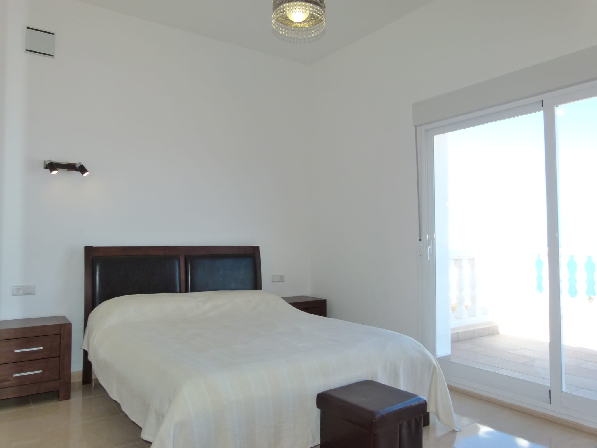 Dormitorio luminoso con cama doble, mesitas de noche de madera oscura y acceso a terraza con barandilla blanca.
