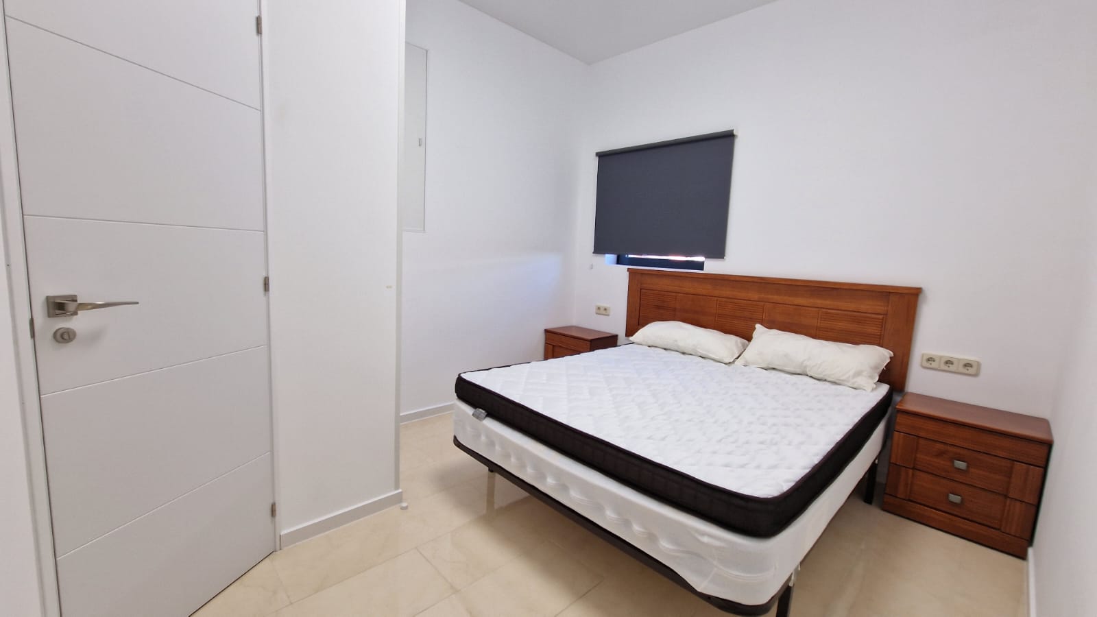 Dormitorio luminoso con cama doble, mesitas de noche de madera y puerta blanca con diseño moderno.