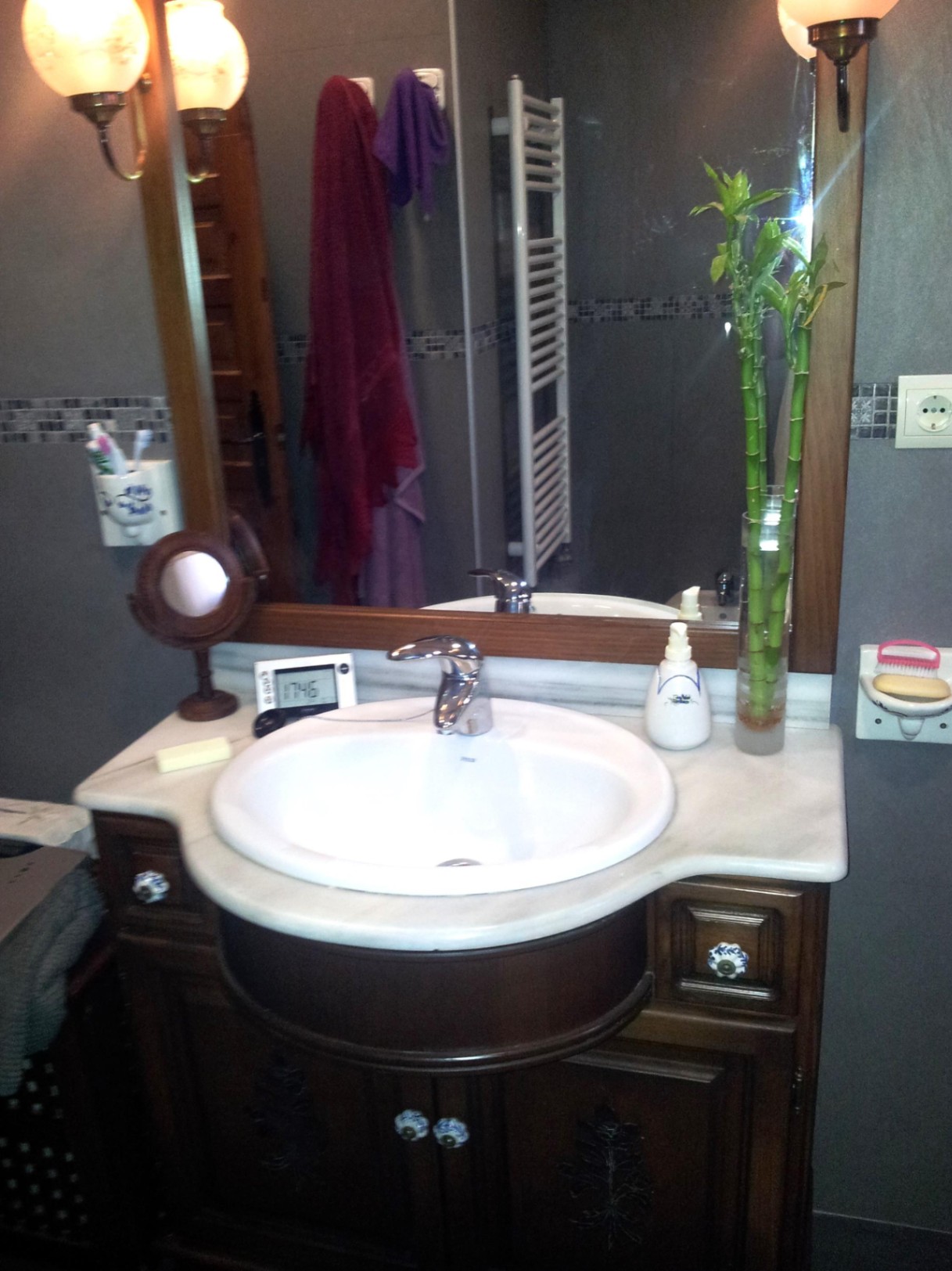 Baño con lavabo encastrado en encimera de mármol blanco sobre mueble de madera oscura. Incluye espejo enmarcado, apliques clásicos y paredes en tonos grises.