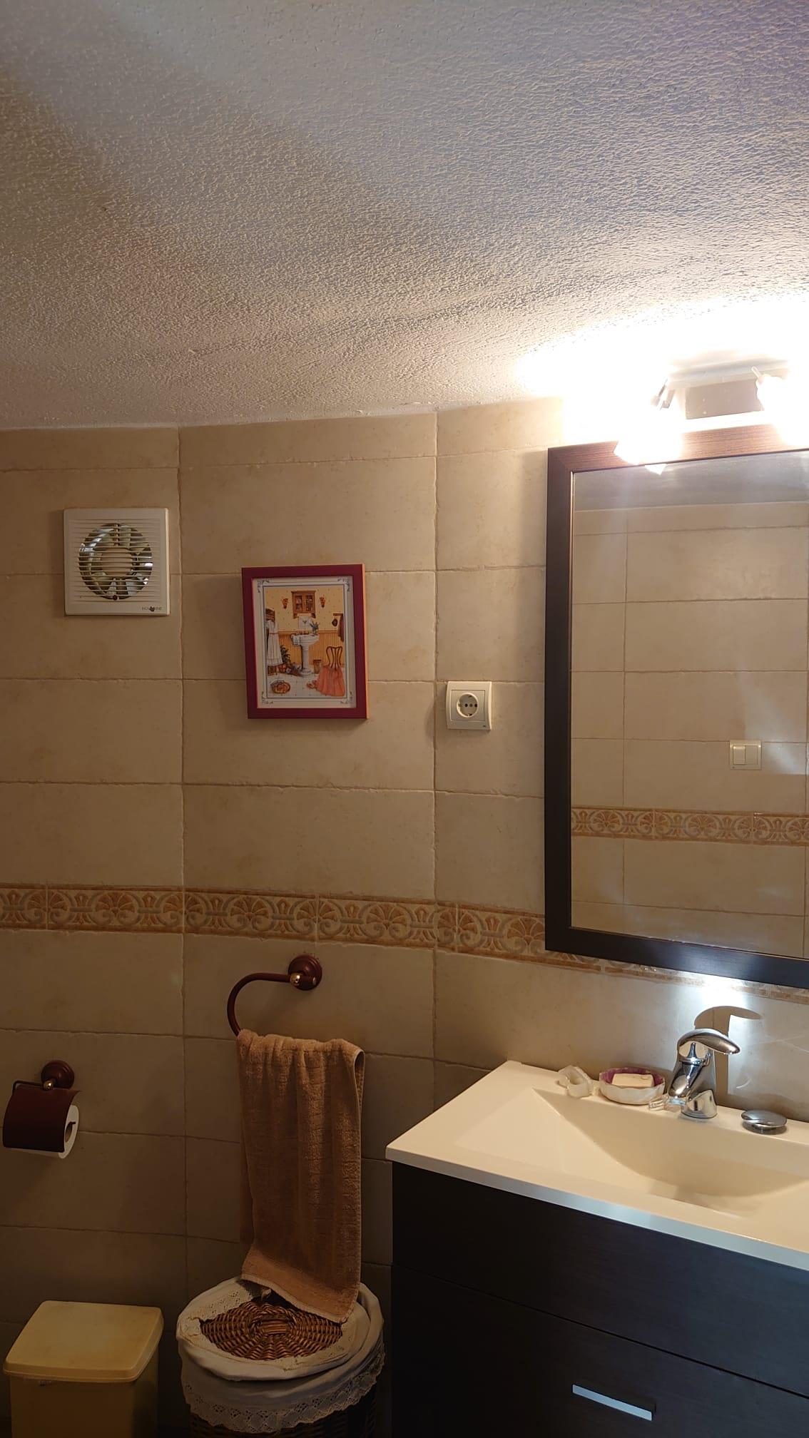 Baño funcional con mueble bajolavabo oscuro, espejo enmarcado y paredes revestidas en azulejos beige con cenefa decorativa y extractor de aire.