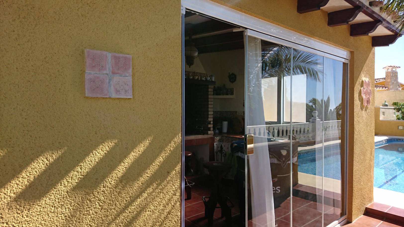 Cocina de verano cerrada con cortinas de cristal, barbacoa de obra y vigas de madera, situada junto a la piscina con suelos de terracota.
