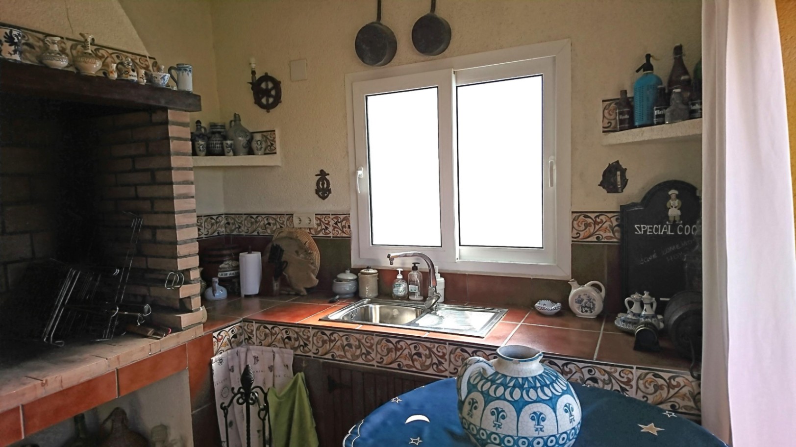 Cocina de verano con barbacoa de obra, encimera de azulejos tradicionales, fregadero de acero inoxidable y ventana de PVC que aporta luz natural.