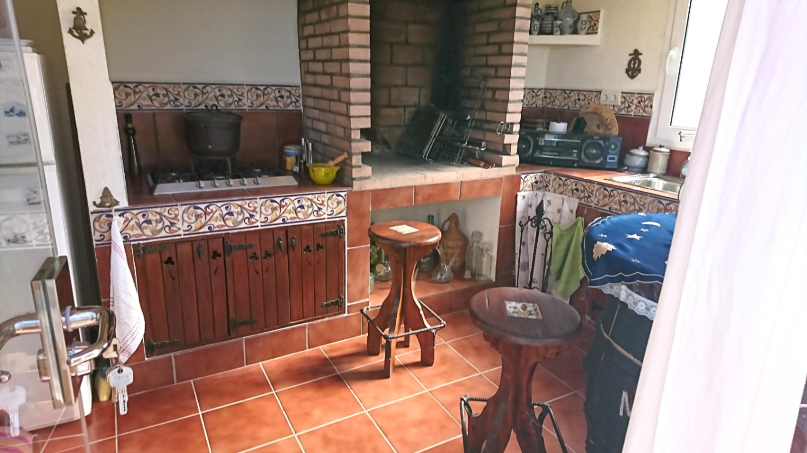 Cocina de verano rústica con barbacoa de obra en ladrillo, encimeras de azulejo decorativo, armarios de madera y suelos de terracota tradicionales.