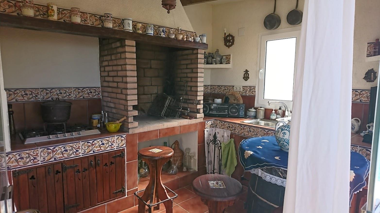 Cocina de verano rústica con barbacoa de obra en ladrillo, encimeras decoradas con azulejos tradicionales, fogones de gas y mobiliario de madera con herrajes clásicos.