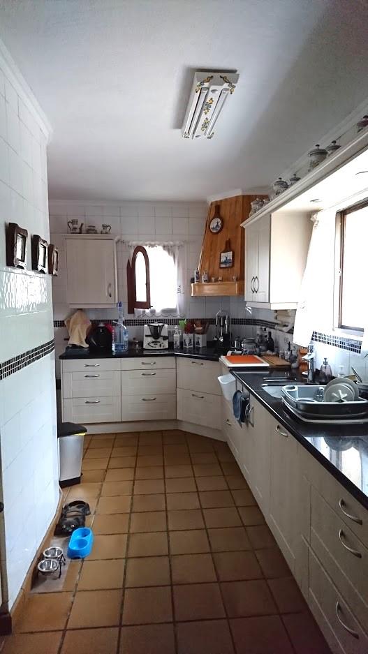 Cocina independiente con mobiliario blanco, encimeras de granito oscuro y suelo de gres. Destaca la campana de madera y la entrada de luz natural por las ventanas laterales.