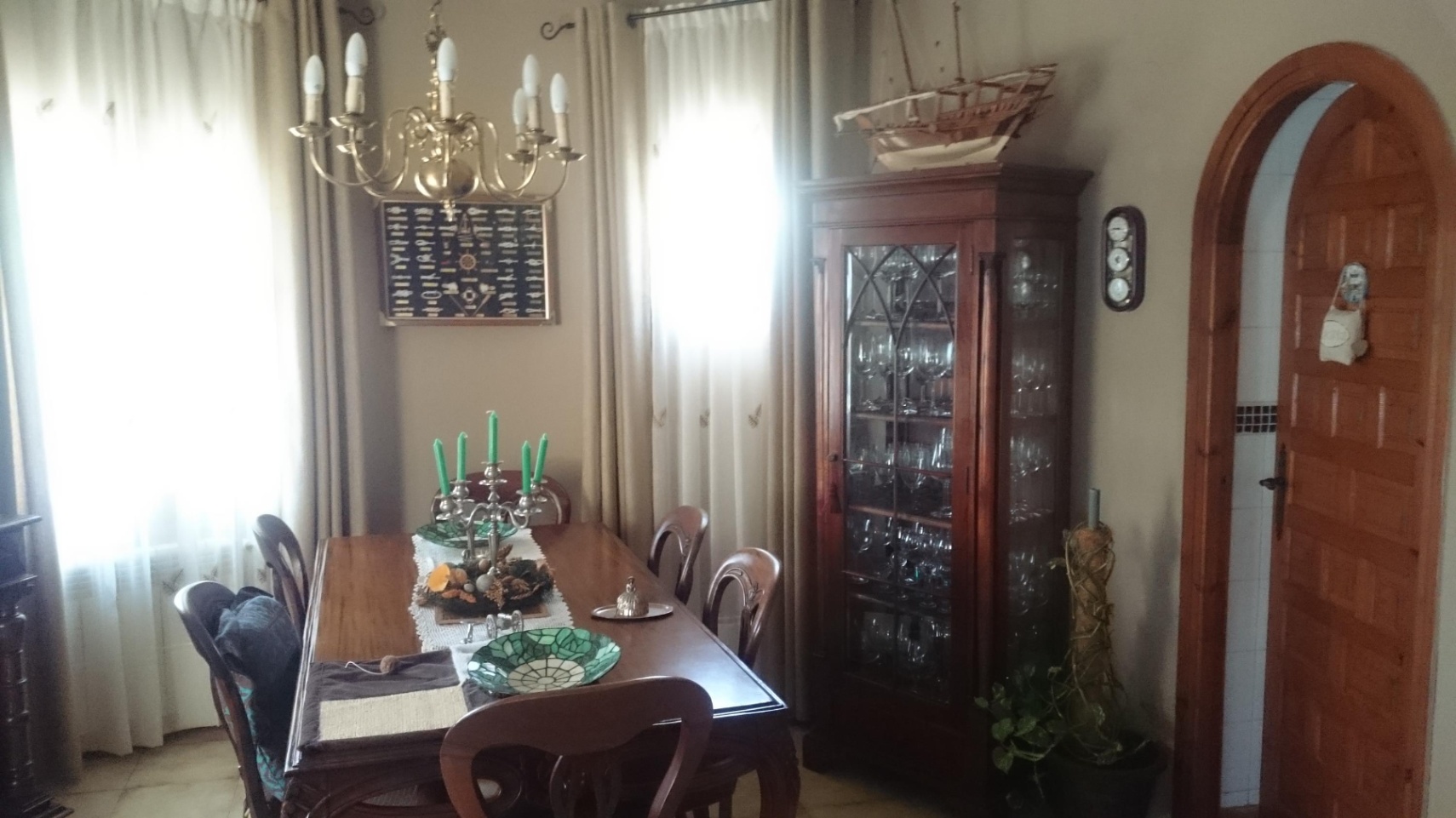 Comedor de estilo clásico con mesa de madera, vitrina acristalada y lámpara de latón. Destaca el arco de madera y la luz natural filtrada por cortinas claras.