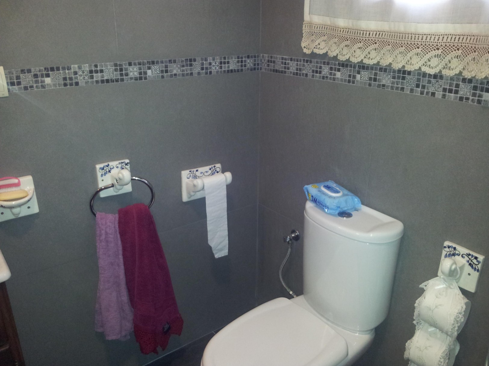 Cuarto de baño con azulejos grises y cenefa de mosaico, equipado con sanitarios blancos y accesorios de cerámica con motivos florales azules.