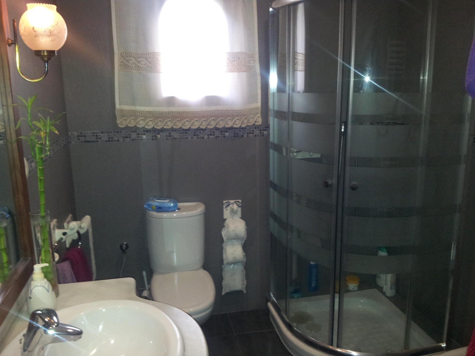 Cuarto de baño con cabina de ducha curva de cristal, paredes en tonos grises con cenefa de mosaico y sanitarios blancos clásicos bajo una luz natural suave.