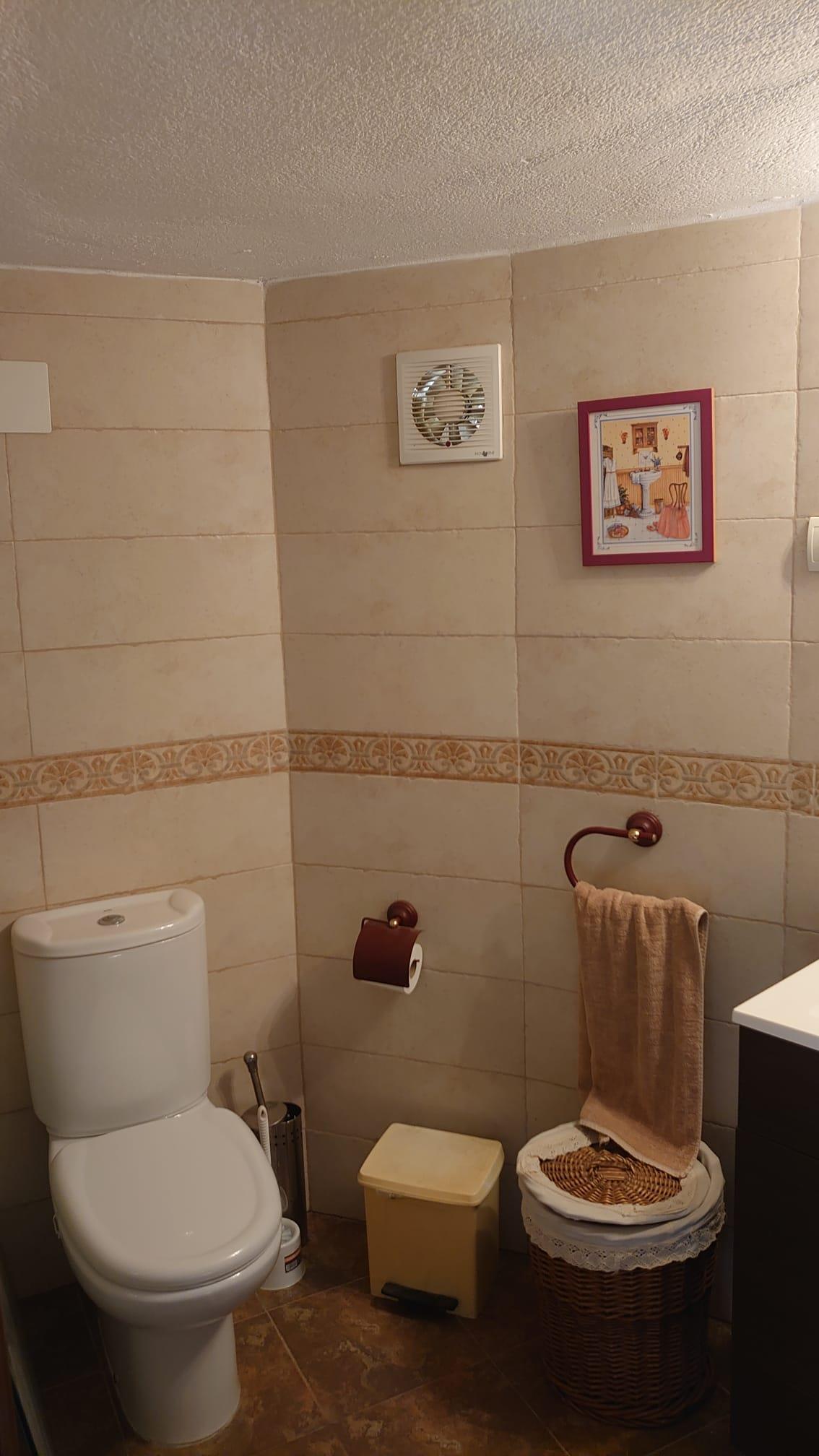 Cuarto de baño con paredes alicatadas en tonos crema y cenefa decorativa. Incluye sanitario de cerámica blanca, accesorios de madera y un cesto de mimbre.