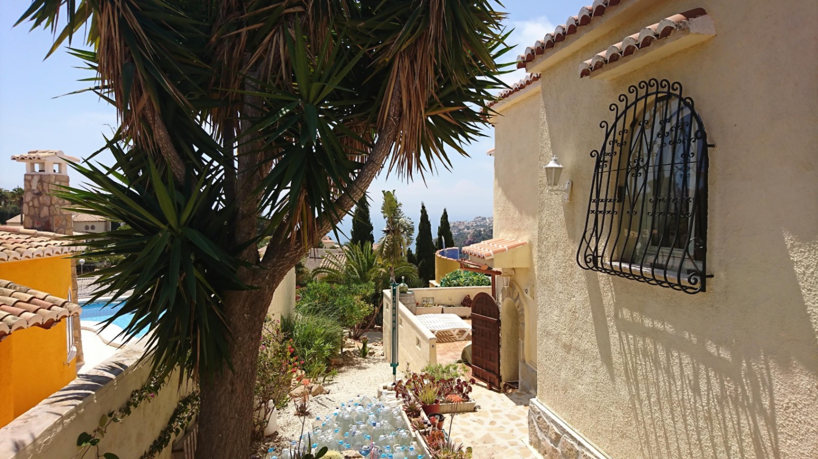 Exterior de la villa con fachada de estuco, ventana con rejería de hierro forjado y jardín mediterráneo con yucas y vistas al mar al fondo.