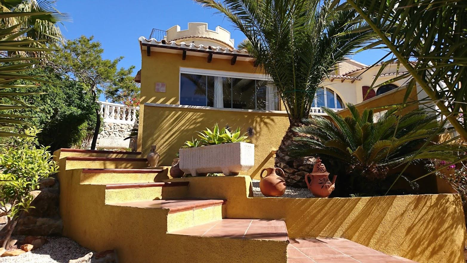 Fachada de color ocre con escaleras de terracota, rodeada de vegetación mediterránea y una torre distintiva con remate almenado bajo el cielo despejado.