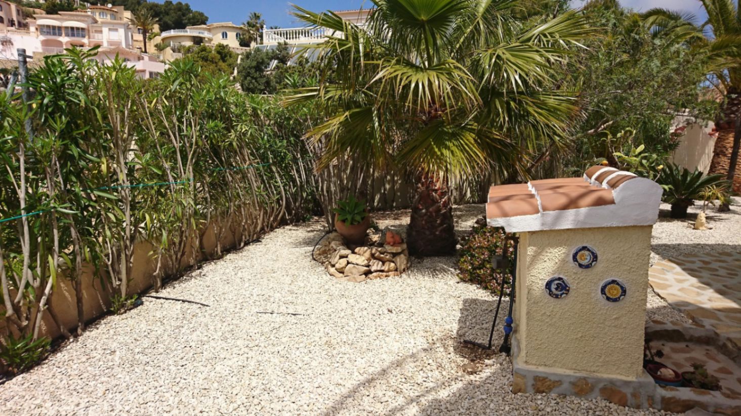Jardín mediterráneo de bajo mantenimiento con grava blanca, palmeras y setos perimetrales, junto a una estructura decorativa con azulejos tradicionales.