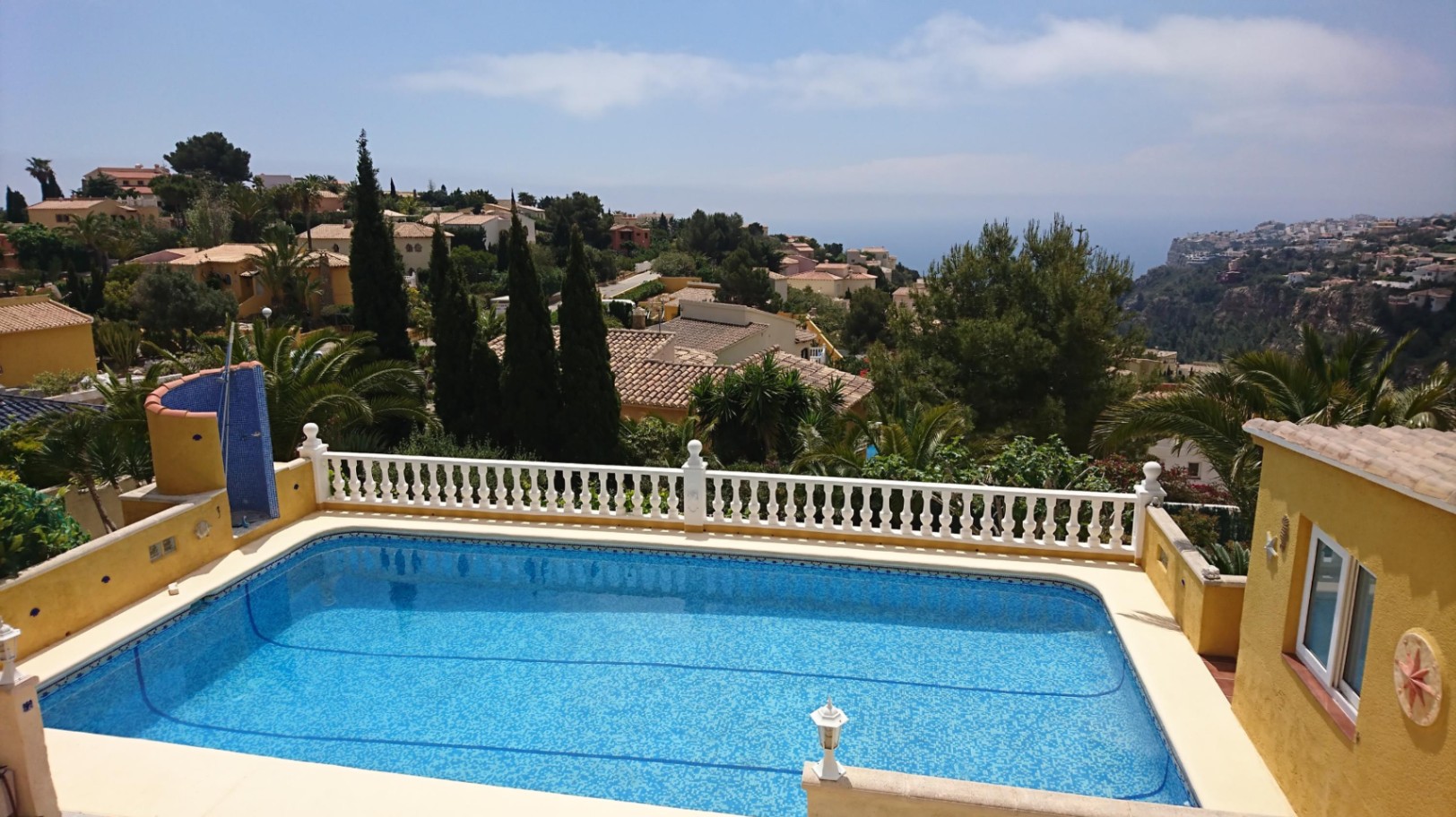 Piscina rectangular con revestimiento de gresite azul, rodeada por una balaustrada blanca clásica y vistas despejadas al mar Mediterráneo y al entorno residencial.