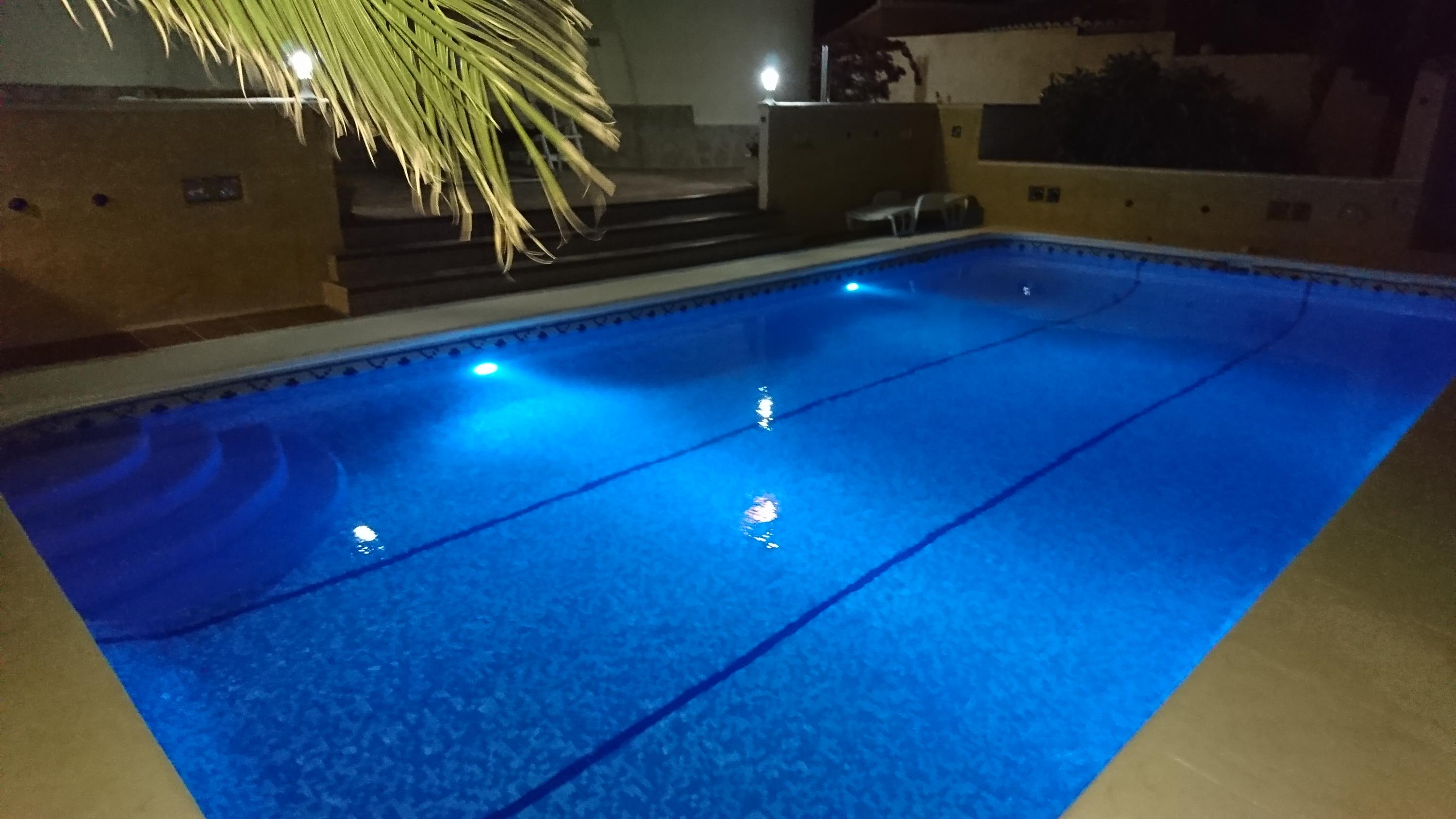Piscina rectangular iluminada con focos subacuáticos azules, rodeada de una terraza pavimentada y muros exteriores con iluminación nocturna.