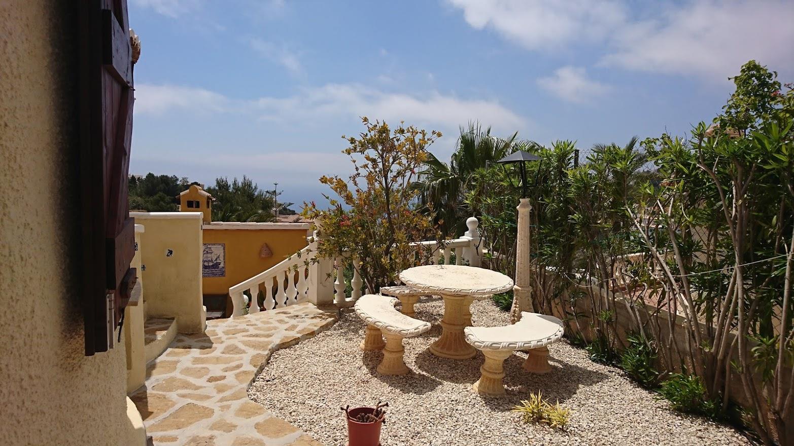 Terraza exterior con mobiliario de piedra tallada sobre suelo de grava, rodeada de vegetación mediterránea y vistas al mar tras la balaustrada blanca.