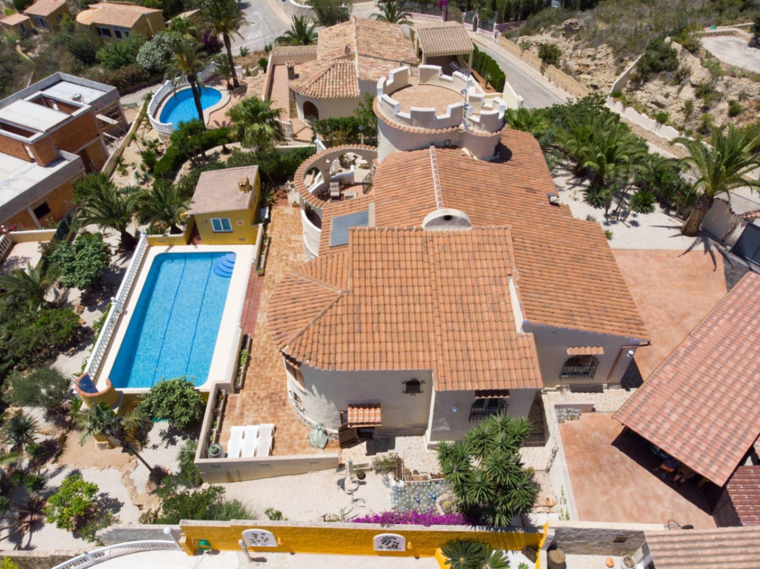 Vista aérea de la villa con tejado de teja cerámica, torreón blanco distintivo, piscina rectangular de 10x4 metros y jardín mediterráneo con palmeras.