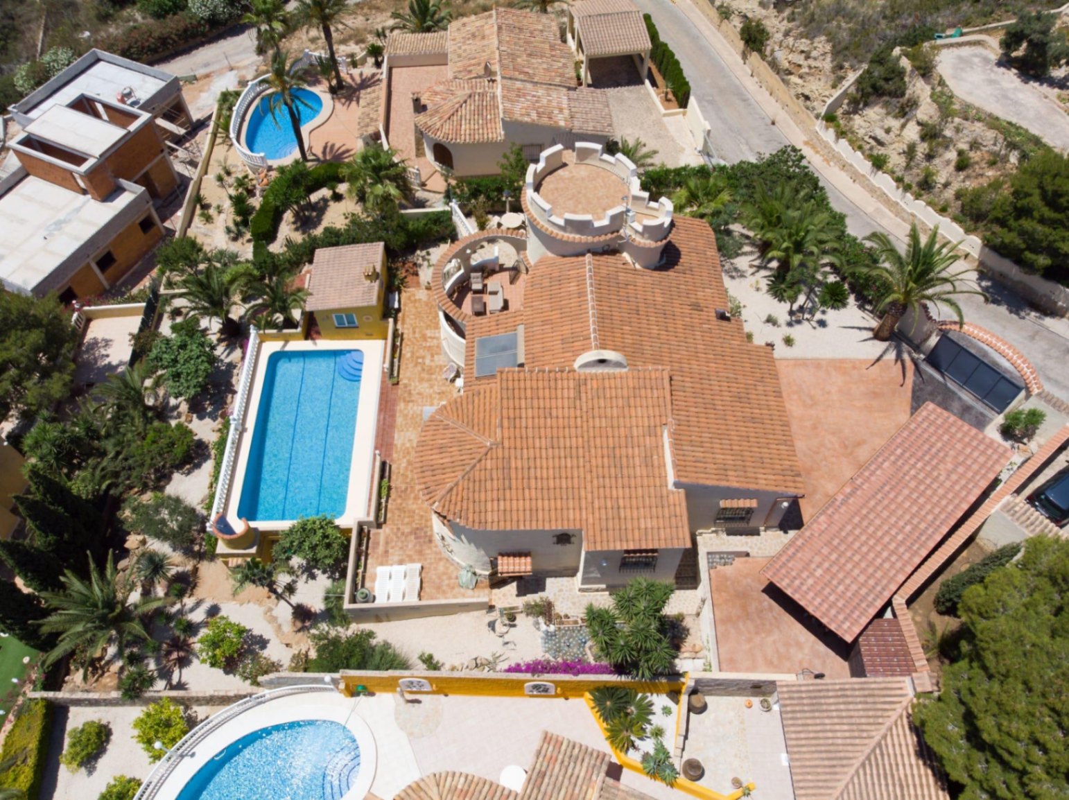Vista aérea de la villa que muestra su tejado de teja cerámica, la característica torre circular con almenas y la piscina rectangular rodeada de vegetación mediterránea.