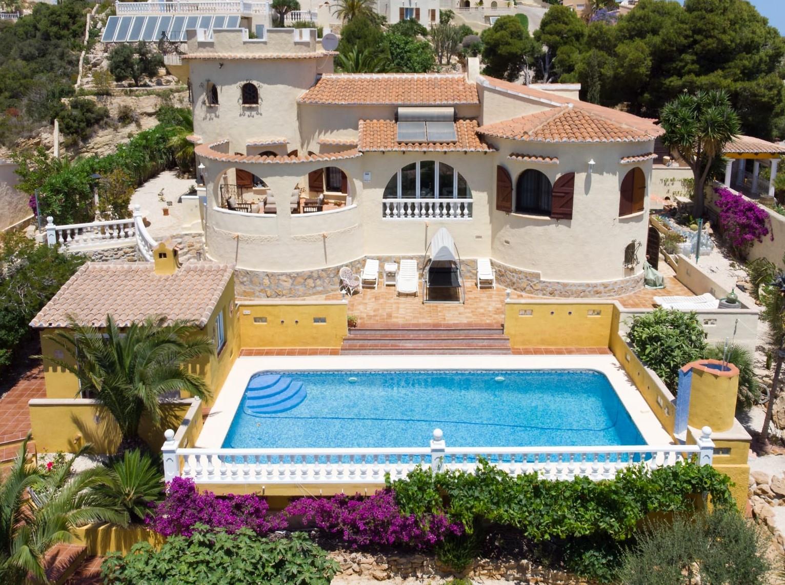 Vista exterior de la villa con piscina rectangular, arquitectura tradicional con torre, tejados de teja y un jardín mediterráneo con buganvillas y palmeras.