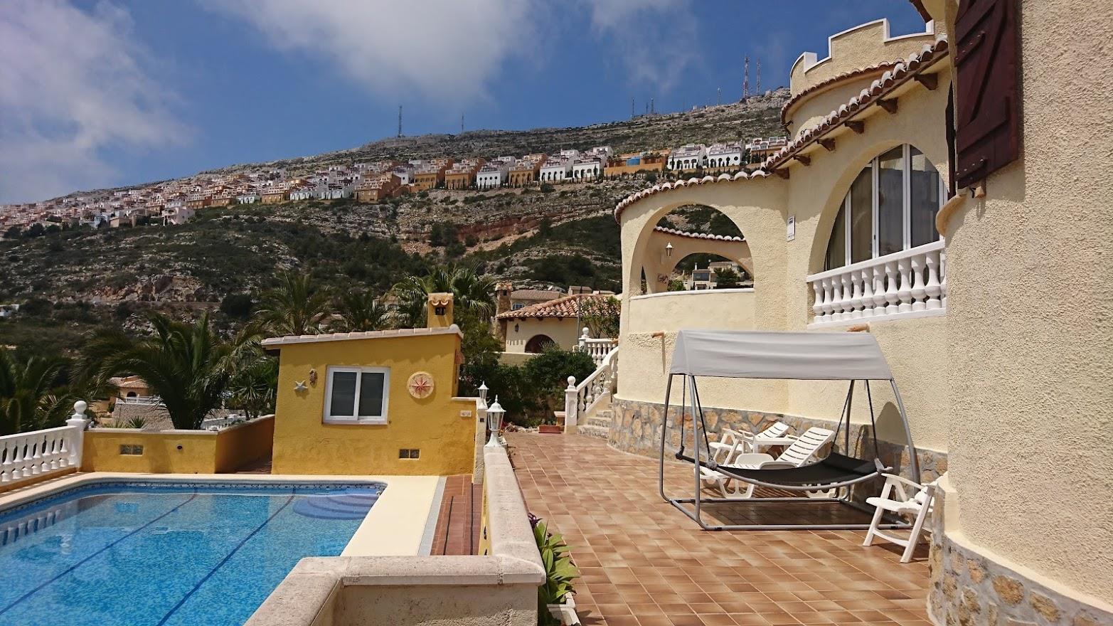 Vista exterior de la villa con piscina, terraza de baldosas y naya arqueada. Al fondo se aprecia la ladera de Cumbre del Sol con su característica urbanización.
