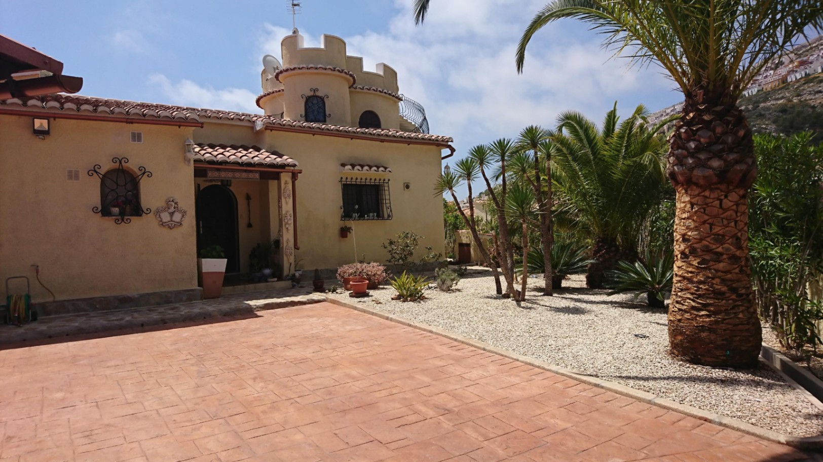 Vista exterior de la villa con su distintiva torre circular, fachada de tonos cálidos y jardín mediterráneo de bajo mantenimiento con palmeras.