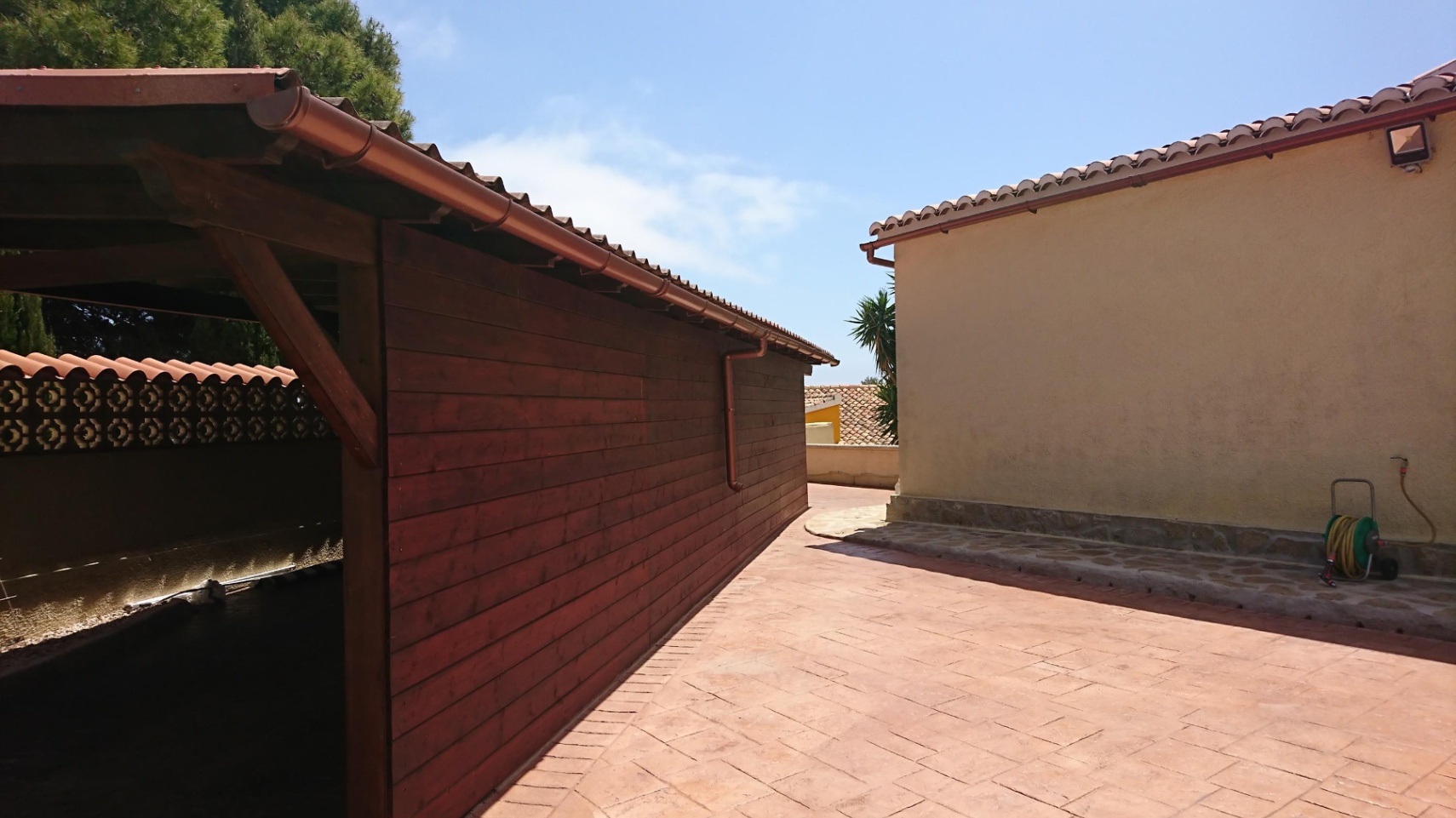 Zona exterior pavimentada con acceso a una estructura de madera para almacenamiento o aparcamiento, junto a la fachada principal con tejados de teja cerámica.