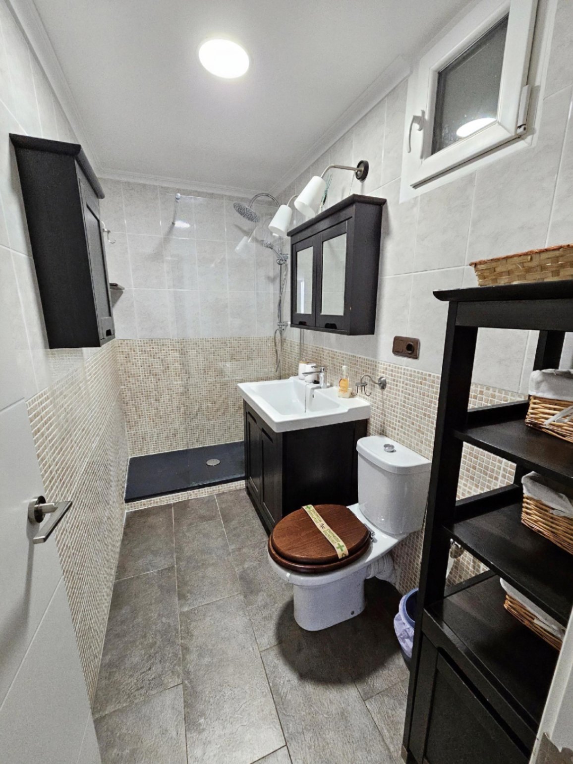 Baño contemporáneo con ducha a ras de suelo, paredes alicatadas con detalle de mosaico, mobiliario de madera oscura y estanterías con cestas de mimbre.