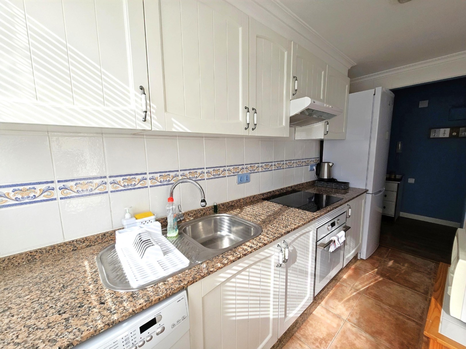 Cocina reformada con encimera de granito, armarios blancos de estilo rústico y salpicadero cerámico decorativo. Incluye placa vitrocerámica y horno encastrado.