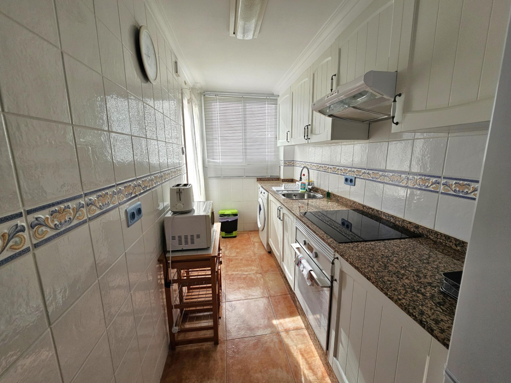 Cocina reformada de estilo alargado con encimera de granito, mobiliario blanco y electrodomésticos integrados. Dispone de luz natural y suelos cerámicos.