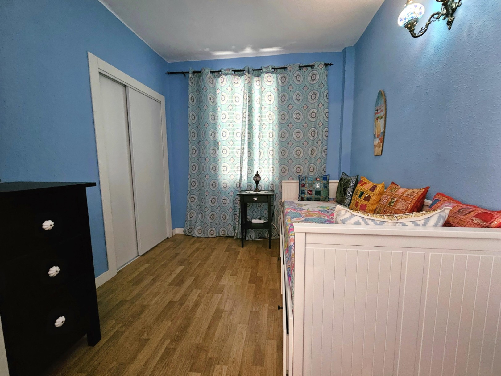 Dormitorio con paredes azul claro, suelo de parqué y armario empotrado blanco. Cuenta con una cama nido blanca, cómoda oscura y textiles de colores vivos.