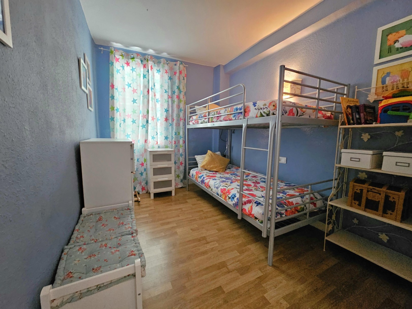 Dormitorio juvenil con literas metálicas, suelos de parqué y paredes en tonos azules. Incluye mobiliario auxiliar blanco y estanterías funcionales con luz natural.